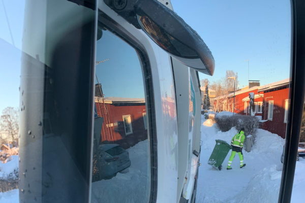 Jäteautonkuljettaja työn parissa näkyy jäteauton sivupeilistä. On talvi.