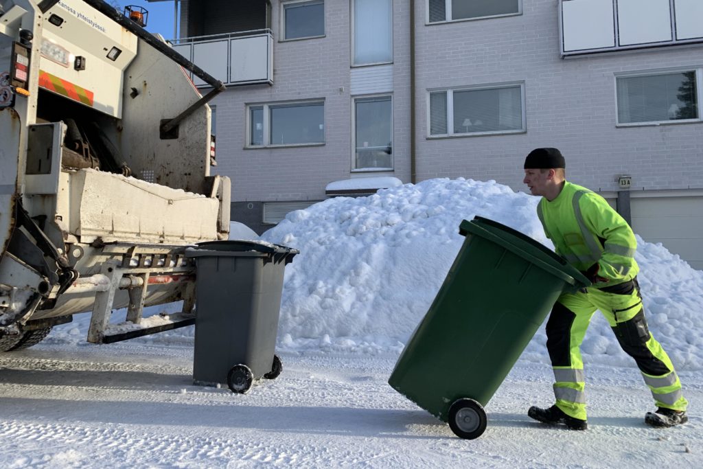 Jäteauton kuljettaja työntää roskista edessään lumisella kadulla. Jäteauton takan odottaa jo toinen jäteastia tyhjennystä.