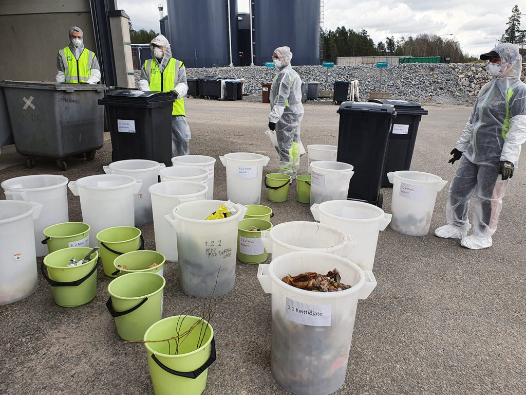Saaveja ja ämpäreitä, joihin on lajiteltu jätteitä sekä tutkimusryhmän jäseniä suojapuvuissa